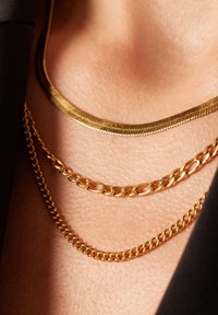 Drie gouden kettingen op de huid gelaagd: een platte slangenketting, een ronde schakelketting en een dikkere curb-ketting, die verschillende texturen tonen.