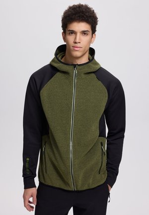 Junger Mann trägt einen grünen, strukturierten Reißverschluss-Hoodie mit schwarzen Ärmeln und seitlichen Reißverschlusstaschen, steht vor einem einfarbigen Hintergrund.