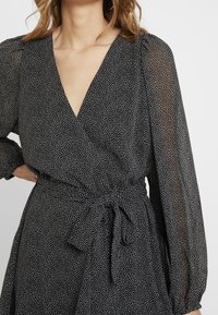 Svart prickigt omslagsklänning med genomskinliga långa ärmar, v-ringning och knytning i midjan. Lätt material med ett flödande textur.