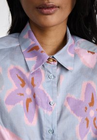 Camicia azzurro chiaro con grande motivo floreale in lavanda e rosa, dotata di colletto button-down e sottile accento dorato sul colletto.