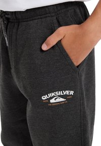 Šedé tepláky s elastickým pásom, predné vrecká a biele a oranžové logo Quiksilver na dolnej časti nohy. Mäkká textúra látky.