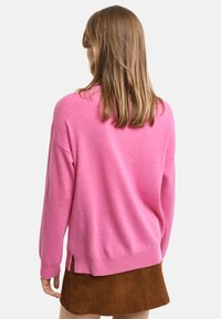 Rosa Strickpullover mit langen Ärmeln, rundem Halsausschnitt und seitlichen Schlitzen. Über einem braunen Wildleder-Minirock getragen. Weiche Textur.