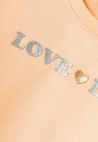 T-shirt couleur pêche avec le mot "LOVE" en paillettes argentées et un accent de cœur doré, présentant une texture douce en coton.