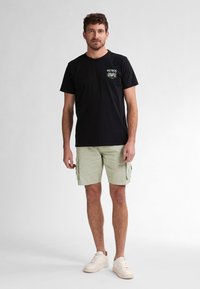 Črna bombažna majica s grafiko "PETROL" na levi strani, v kombinaciji s bež cargo kratkimi hlačami in off-white supergami. Enostavna ozadje.