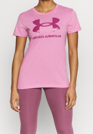 Kvinde iført lyserød Under Armour t-shirt og mauve leggings, står med arme afslappet ned langs siden mod en ensfarvet baggrund.