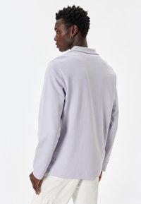 Camisa polo de manga larga en un suave color púrpura claro, con una textura suave, corte relajado y un diseño clásico de cuello. Minimalista, sin patrones visibles ni herrajes.