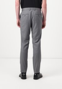 Graue maßgeschneiderte Hose mit einem dezenten Karomuster, ausgestattet mit zwei Gesäßtaschen und einem schlanken Schnitt. Getragen mit schwarzen Schuhen.