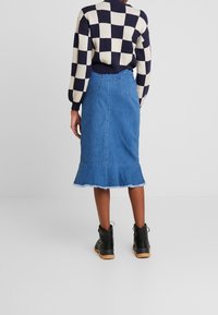 Jupe en denim avec un ourlet effiloché, de couleur bleu moyen, et deux poches arrière. Associée à une bottine noire et un pull à carreaux à motifs.