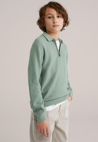 Garçon aux cheveux bruns bouclés portant un pull vert clair texturé à demi-fermeture éclair et un pantalon beige, debout avec une main dans la poche.