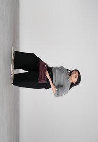 Vrouw in een grijze trui, zwarte wijde broek, witte hoge hakken, met een bordeauxrode clutch, staand op een grijze vloer.