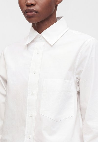 Personne portant une chemise blanche boutonnée avec un col pointu et une poche poitrine, visible des épaules jusqu'au milieu du torse.