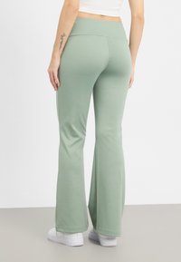 Pantalons évasés en tissu vert sauge doux, avec une large ceinture et une texture lisse, associés à des baskets blanches.