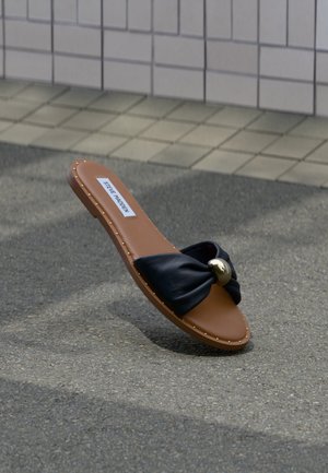 Bruine platte sandaal met zwarte gedraaide band en gouden ronde accent, gelabeld Steve Madden, rustend op een grijze bestrating nabij een betegelde muur.