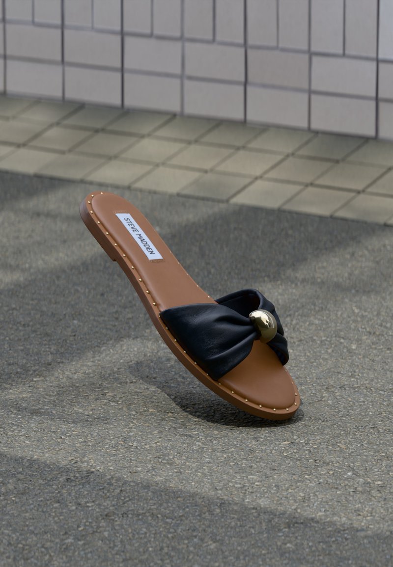 Sandale plate marron avec une lanière noire torsadée et un accent rond doré, étiquetée Steve Madden, reposant sur un trottoir gris près d’un mur carrelé.
