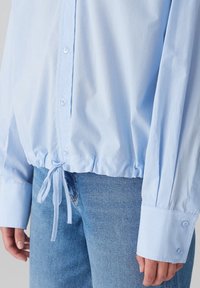 Lyseblå skjorte med knapper, snøre i kanten, lange ærmer og manchetter med knap. Fremstillet af glat, let stof.