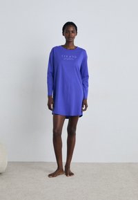 Vestido longo roxo de mangas compridas com um corte descontraído, feito de tecido suave. Apresenta um gráfico da fase da lua em branco na frente. Modelo descalço em pé sobre um tapete bege.