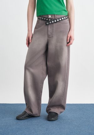 Isik, kes kannab laiuvaid halli pükse, rohelist särki, musta vööd hõbedaste aukudega ja musti tossusid ning seisab sinisel vaibal.