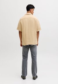 Mees seisab seljaga kaamera poole, kandes beeži lühikeste varrukatega tekstuuriga särki, halli teksapükse ja musti tenniseid tavalise valge tausta ees.