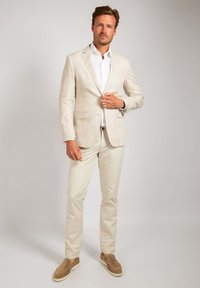 Linnen blazer in lichtbeige, gecombineerd met een witte shirt en crèmekleurige broek. Bruine suède instappers maken de outfit compleet.