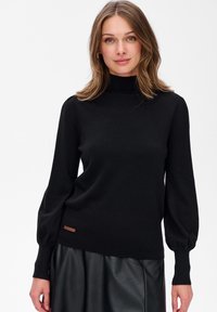 Frau mit schulterlangem Haar, die einen schwarzen Rollkragenpullover und einen schwarzen Lederrock trägt und vor einem weißen Hintergrund steht.