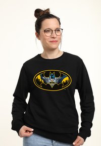 Felpa nera con un grande logo di Batman giallo e grigio accompagnato da un'illustrazione del personaggio. Il tessuto sembra morbido, con collo a giro e maniche lunghe.