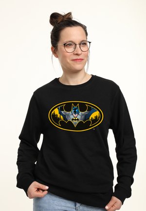 Sweatshirt noir avec un grand logo Batman jaune et gris accompagné d'une illustration du personnage. Le tissu semble doux, avec un col rond et des manches longues.