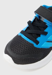 Športni čevlji iz črnega mrežastega materiala, z modrimi poudarki in zaponko s trakom na ježka, črnim vezalkami ter teksturirano belo gumijasto podplatom.