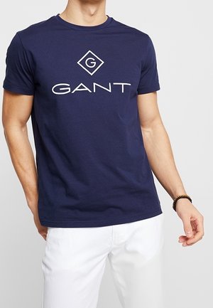 T-shirt en coton bleu marine avec un grand logo "GANT" blanc et un motif diamant. Manches courtes et col rond, présenté avec un pantalon blanc.