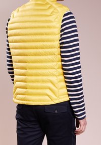 Doudoune jaune, effet gonflé, avec un design matelassé horizontal, en matériau léger, col montant, portée par-dessus une chemise à manches longues rayée bleu marine et blanche.