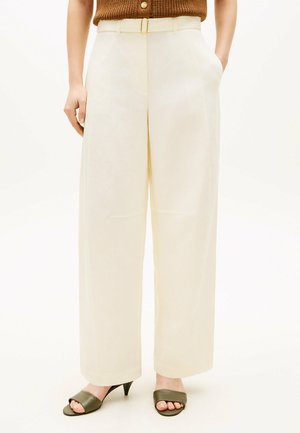 Donna che indossa pantaloni a gamba larga color crema a vita alta con una cintura sottile, abbinati a sandali con tacco aperti verdi oliva e un top marrone senza maniche.