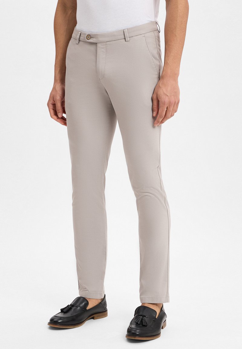 Pantaloni tan slim-fit realizzati in tessuto liscio, con una parte frontale piatta, tasche laterali e chiusura con un solo bottone. Indossati con mocassini neri.
