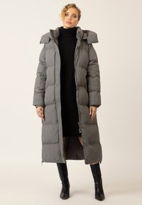 Langer grauer Daunenmantel mit Kapuze, mit einem Reißverschluss auf der Vorderseite, gestepptem Design und Seitentaschen. Getragen über einem schwarzen Strickkleid und kniehohen Stiefeln.