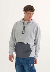 Sudadera con capucha gris que tiene un cordón negro y un gran bolsillo gris en la parte frontal. Las telas parecen suaves con un corte relajado.
