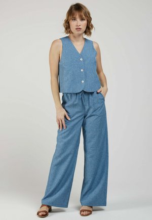 Femme debout, portant un gilet bleu sans manches boutonné, un pantalon large assorti et des sandales marron à bouts ouverts sur fond uni.