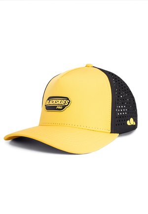 Junge Frau mit lockigem dunklem Haar, die eine gelbe und schwarze "BLACKSKIES PRO" Cap trägt und leicht seitlich vor neutralem Hintergrund schaut.
