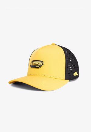 Ung kvinde med krøllet mørkt hår iført en gul og sort "BLACKSKIES PRO" cap, der kigger let til siden mod en neutral baggrund.
