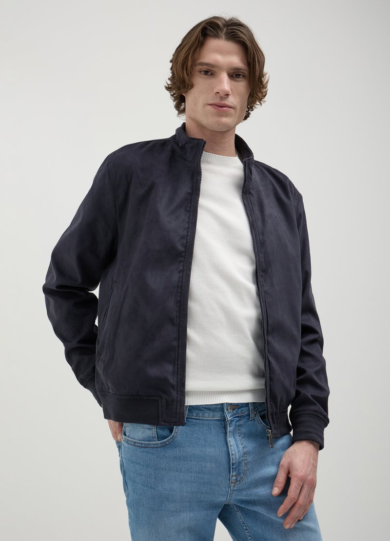 Veste bomber bleu marine foncé en tissu lisse, dotée d'un col montant, d'une fermeture éclair et de poches latérales, portée par-dessus un pull léger et un jean bleu.