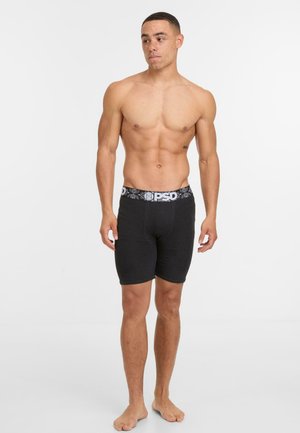 Shirtless man with freckles wearing black PSD boxer briefs with a black and white waistband, standing barefoot on a white background.