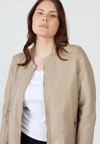Beige läderjacka med rund krage, framdragkedja och sidfickor. Bärs över en vit topp, har en slät yta och figursydd design.