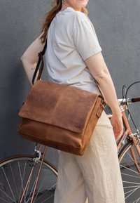 Brązowa skórzana torba listonoszka z klapą, prostokątnym kształtem i regulowanym paskiem. Charakteryzuje się gładką fakturą oraz subtelnym logo.