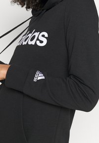Personne portant un sweat à capuche Adidas noir avec un logo blanc sur la poitrine et la manche, tenant un cordon noir près du cou.