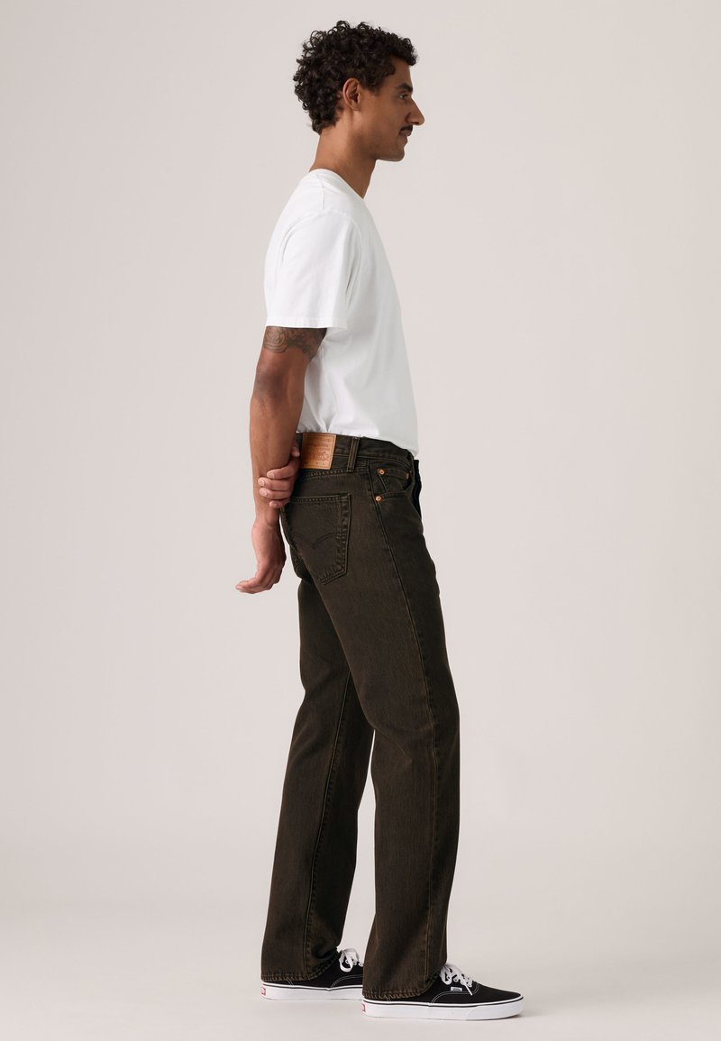 Mand i hvid t-shirt, mørke jeans og sorte sneakers står sidelæns med hænderne bag ryggen mod en ensfarvet lys baggrund.