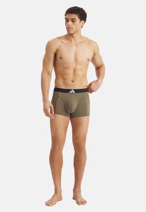 Athletic man standing barefoot wearing olive green boxer briefs with a black waistband, looking to his left with one hand on hip.