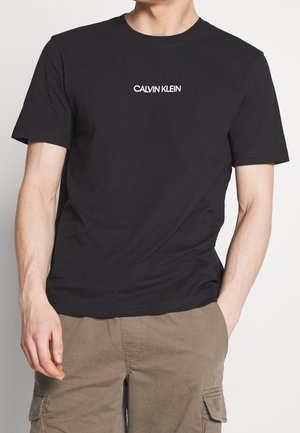 Czarna bawełniana koszulka z krótkim rękawem, z białym logo "CALVIN KLEIN" na klatce piersiowej. Okrągły dekolt, gładka tekstura.