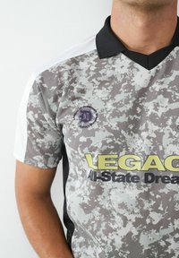 Koszulka polo z krótkim rękawem w szarym wzorze moro z czarnym kołnierzykiem, z napisem "LEGACY All-State Dreams" w żółtym i czarnym kolorze oraz haftowanym logo.