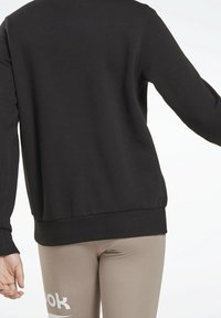 Sudadera negra de manga larga, con puños y bajo acanalados, con una textura suave. Combinada con leggings beige, logo de la marca visible.