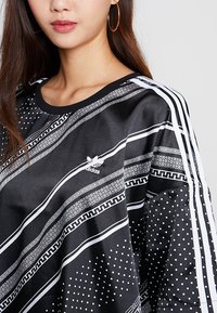 Svart idrottströja med vita geometriska mönster, polka punkter och tre ränder längs ärmarna. Adidas-logotypen på bröstet.