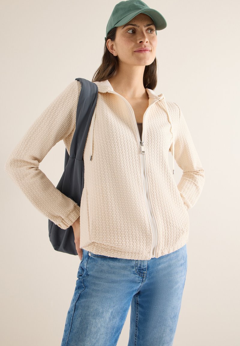 Beige gestructureerd vest met rits, capuchon en trekkoord, gecombineerd met een blauwe spijkerbroek en een grijze totebag. Model draagt een groene pet.
