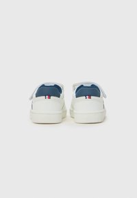 Baskets blanches avec un talon bleu et une semelle blanche texturée. L'arrière présente un détail de logo minimaliste rouge, blanc et bleu.