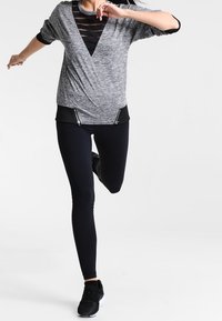 Top atletico grigio leggero con scollo a V, inserti in rete nera e fondo nero a contrasto. Abbinato a leggings neri e scarpe da ginnastica.